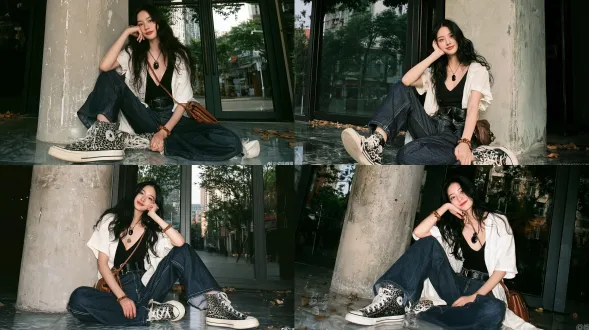 全身镜头，年轻貌美的东亚女性，黑色长波浪卷发，温婉浅笑，随意坐在城市街边店铺门口的水磨石地面上，倚靠在饱经风化的粗糙混凝土立柱上；身着宽松敞开的白色短袖衬衫，内搭黑色深v吊带，深蓝色阔腿牛仔裤搭配黑色皮质腰带，黑白豹纹高帮帆布鞋；脖颈佩戴复古黑玛瑙吊坠项链，手腕戴着木珠手链，棕色皮质单肩包斜挎肩头；双腿分开弯曲摆出姿势，左手轻触脸颊，右手搭在膝盖上；背景是巨大的落地玻璃门，玻璃映照出街边树木与远处城市楼宇，地面散落少许落叶；自然柔和侧光，复古柯达金200胶片质感，细腻胶片颗粒，低饱和冷色调，随性倾斜构图，自然浅景深，背景虚化，纪实街头摄影氛围。避免 ai 美女脸、低质量、模糊、肢体扭曲、多余肢体、多余手指、畸形特征、曝光过度、曝光不足、噪点、粗糙皮肤、肤色暗沉、表情僵硬、过度平滑、塑料质感、水印、文字、边框、过度精修、塑料皮肤、暗沉肤色、明显痘印、明显皱纹、斑点、面部变形、肢体变形。