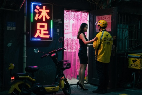 街拍，夜晚，城中村，一个暗黑的门口被深粉色蕾丝半透明窗帘遮住，里面光线昏暗，边上站着1个25岁女人，穿着包臀裙，黑色高跟鞋，搂着一个黄色服装的外卖小哥就要进屋，头上的霓虹灯广告牌文字：“沐足”,前景是一辆美团电瓶